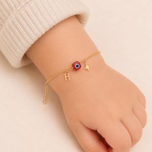 Op de afbeelding: Een goudkleurige kettingarmband met een kleine rode vierkante kraal met een blauw-wit oogmotief, een goudkleurige letter H-bedel en een goudkleurige sterbedel. De fijne metalen ketting is voorzien van een karabijnsluiting en een verlengstuk.