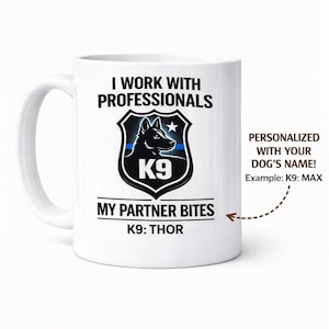 May include: White ceramic mug with a K9 design. The mug features the text "I WORK WITH PROFESSIONALS" and "MY PARTNER BITES". A black and white dog graphic is in the center. The mug is personalized with the dog's name.