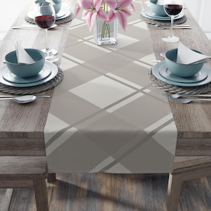 May include: A neutral-toned table runner with a geometric plaid pattern. The runner features a repeating design of intersecting lines and squares in shades of gray and white. The table is set with teal-colored bowls, plates, and wine glasses, creating a coordinated dining setting.