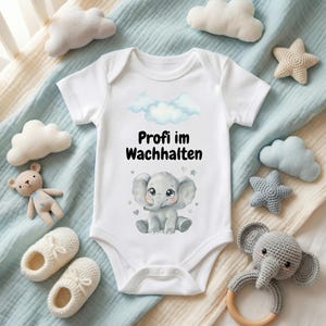 Könnte beinhalten: Weißer Baby-Body mit dem Text "Profi im Wachhalten" und einer Elefanten-Illustration. Das Bild zeigt Baby-Accessoires wie wolkenförmige Kissen, einen Teddybär und Babyschuhe, auf einer hellblauen Decke.