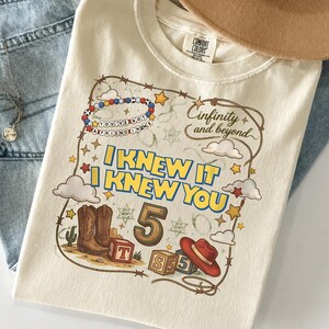 May include: A cream-colored t-shirt featuring a graphic print with the text "I KNEW IT I KNEW YOU", "infinity and beyond", and "YOU'VE GOT A FRIEND IN ME". The design includes a cowboy boot, a red hat, blocks with letters, the number 5, and stars.