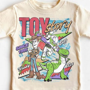 May include: Cream-colored t-shirt featuring the words "TOY STORY" in red and the phrase "SINCE 1995." The shirt has a graphic of Woody, Buzz Lightyear, and Rex the dinosaur. The shirt is a children's apparel item.
