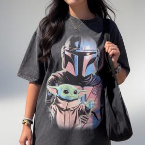 May include: Dark gray t-shirt featuring a graphic of The Mandalorian and Grogu. The design includes the Mandalorian's helmet in blue and pink, and Grogu in a pink and white outfit. The shirt has short sleeves and a relaxed fit.