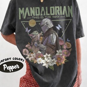 May include: Dark gray t-shirt featuring a graphic of The Mandalorian and Grogu surrounded by flowers and butterflies. The text "MANDALORIAN - THIS IS THE WAY" is printed above the image. The shirt has a Comfort Colors Pepper label.