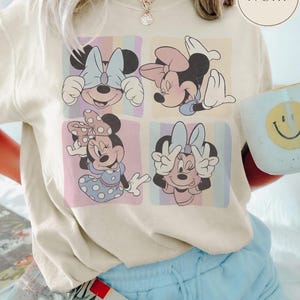 May include: Cream-coloured t-shirt with a four-panel Minnie Mouse graphic. Each panel displays a different pose of Minnie Mouse against a pastel striped backdrop. The shirt is worn with light blue shorts.