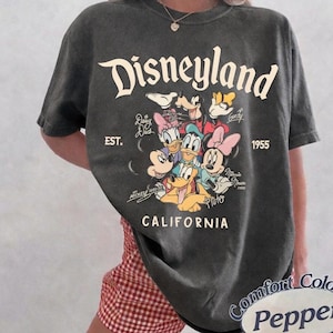 May include: Dark gray t-shirt featuring the Disneyland logo and various Disney characters. The shirt includes the text "EST. 1955" and "CALIFORNIA". The shirt is a Comfort Colors Pepper brand.