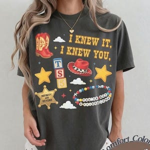 May include: A charcoal gray short-sleeve t-shirt featuring a graphic print with the text I KNEW IT, I KNEW YOU, a red cowboy boot, a red cowboy hat, yellow stars, a sheriff badge with INFINITY AND BEYOND, alphabet blocks, and a beaded bracelet.