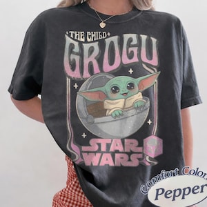 Puede incluir: Camiseta gris oscuro con Grogu de Star Wars. El dise&ntilde;o incluye el texto "THE CHILD + GROGU" en una fuente retro, con Grogu en su c&aacute;psula. El logotipo de Star Wars tambi&eacute;n es visible.