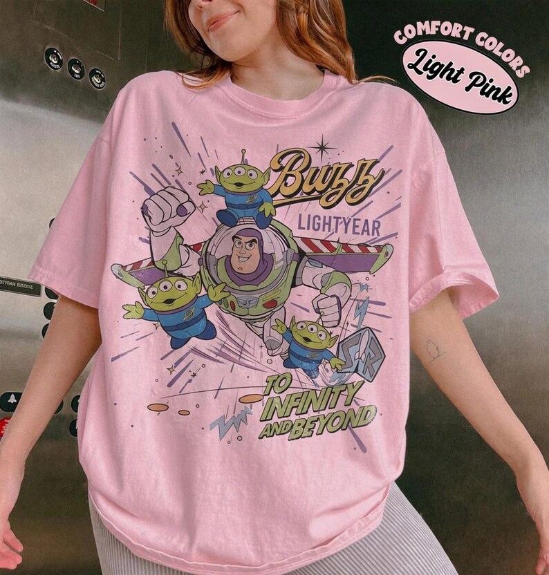 May include: Light pink t-shirt featuring Buzz Lightyear and the aliens from Toy Story. The graphic includes the words "Buzz Lightyear" and "To Infinity and Beyond". The shirt has a relaxed fit and the words "Comfort Colors Light Pink" are printed on the top right.