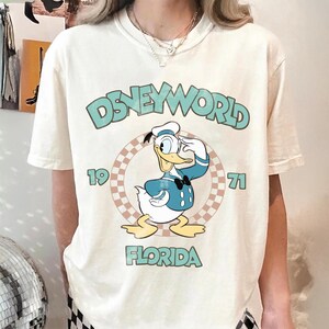May include: Cream-colored t-shirt featuring a graphic of Donald Duck saluting, set within a checkered circle. The text "DISNEYWORLD 1971 FLORIDA" is printed above and below the graphic in a teal font. The shirt has short sleeves.