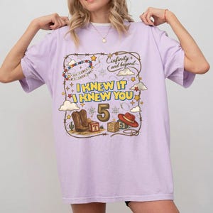 May include: A light purple t-shirt featuring a graphic print with the text "I KNEW IT I KNEW YOU" and "Cinfinity and beyond". The design includes a cowboy hat, boots, alphabet blocks, stars, clouds, and a beaded string with the text "YOU'VE GOT A FRIEND IN ME".