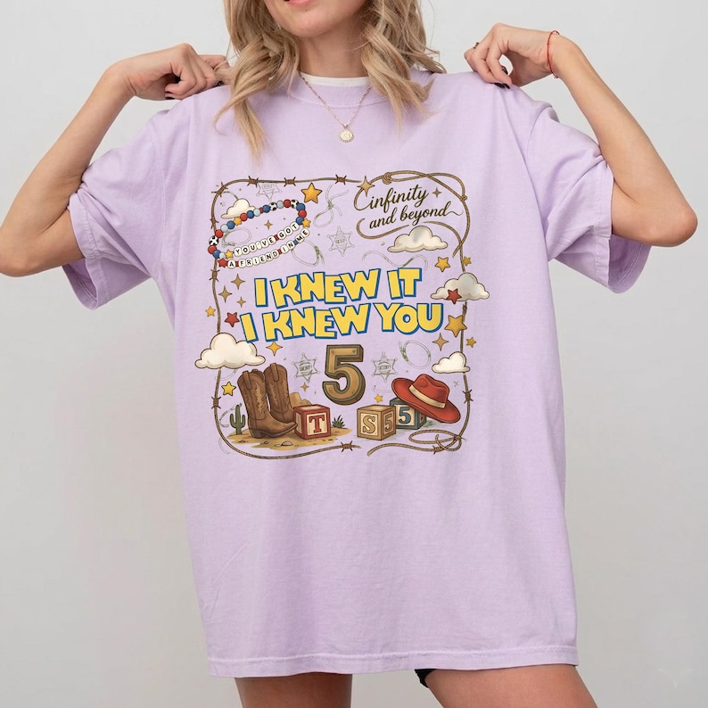 May include: A light purple t-shirt featuring a graphic print with the text "I KNEW IT I KNEW YOU" and "Cinfinity and beyond". The design includes a cowboy hat, boots, alphabet blocks, stars, clouds, and a beaded string with the text "YOU'VE GOT A FRIEND IN ME".