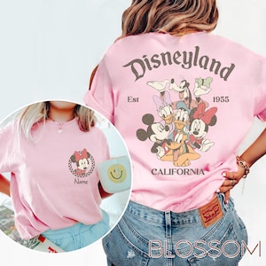 May include: Pink t-shirt featuring a Minnie Mouse graphic on the front and a Disneyland design on the back. The Disneyland design includes Mickey Mouse, Donald Duck, Goofy, and other characters. The shirt has a vintage, retro style.