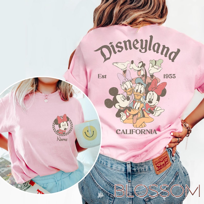 May include: Pink t-shirt featuring a Minnie Mouse graphic on the front and a Disneyland design on the back. The Disneyland design includes Mickey Mouse, Donald Duck, Goofy, and other characters. The shirt has a vintage, retro style.