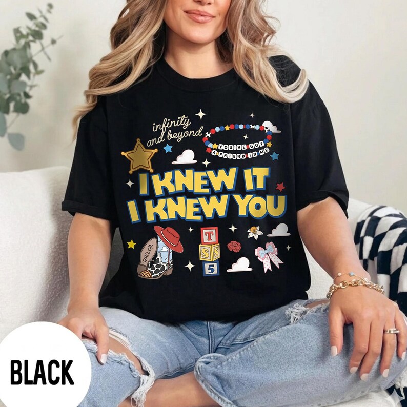 May include: A black short-sleeve t-shirt featuring a graphic print with the text "infinity and beyond", "YOU'VE GOT A FRIEND IN ME", "I KNEW IT", and "I KNEW YOU". The design includes a gold star, a beaded bracelet, a cowboy boot, and alphabet blocks.