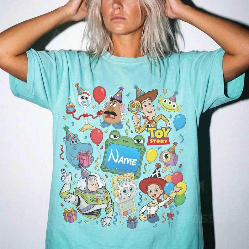 May include: A light turquoise t-shirt featuring a colorful Toy Story graphic print. The design includes characters like Woody, Buzz Lightyear, Jessie, and Mr. Potato Head wearing party hats, surrounded by balloons, gifts, and a frog-shaped frame with the text Name.