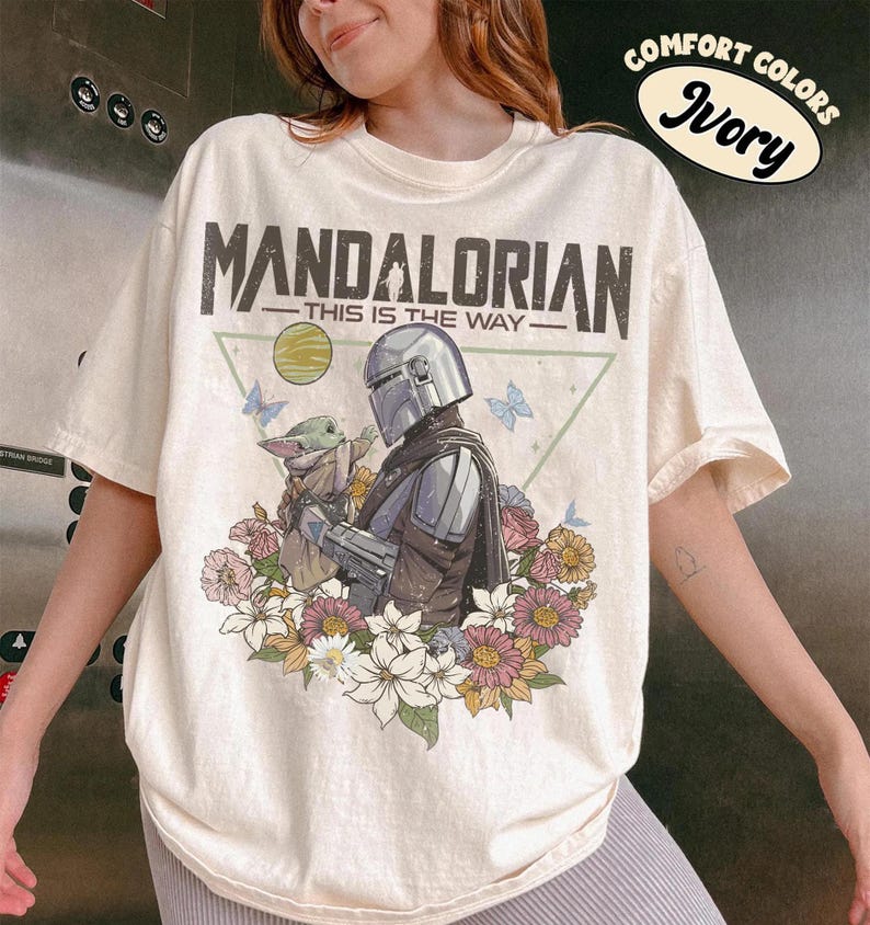 May include: Ivory-colored t-shirt featuring a graphic of The Mandalorian and Grogu surrounded by flowers and butterflies. The text "MANDALORIAN - THIS IS THE WAY" is printed above the image. The shirt has short sleeves and a crew neck.
