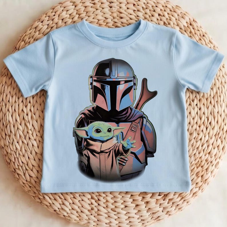 K&ouml;nnte beinhalten: Ein hellblaues Kurzarm-T-Shirt mit einem Grafikdruck eines behelmten Charakters in dunkler R&uuml;stung, der hinter einer kleinen gr&uuml;nen Kreatur mit gro&szlig;en Ohren und Augen steht, alles im Stil einer illustrierten Comic-Grafik dargestellt.