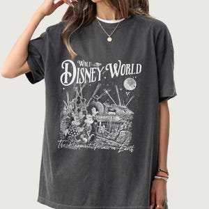 May include: Dark gray t-shirt featuring a black and white graphic of Mickey and Minnie Mouse in front of a Disney World scene. The text "Walt Disney World" and "The Happiest Place on Earth" are also displayed on the shirt.