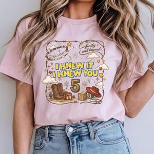May include: A light pink t-shirt featuring a graphic print with a lasso border, cowboy boots, a red hat, alphabet blocks, and stars. The text reads "Cinfinity and beyond", "I KNEW IT I KNEW YOU", and "YOU'VE GOT A FRIEND IN ME" with the number 5.