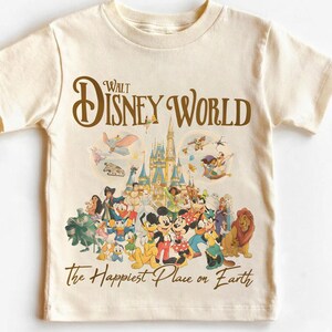 May include: Cream-colored t-shirt featuring a graphic of Disney World characters and the text "Walt Disney World" in brown. The shirt also includes the phrase "The Happiest Place on Earth" in a script font.