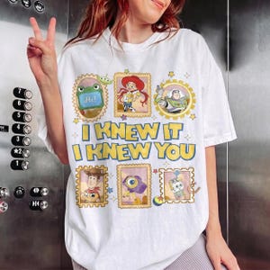 May include: A white short-sleeve t-shirt featuring a graphic print of Toy Story characters in postage stamp frames. The text reads I KNEW IT I KNEW YOU in yellow with blue outlines. Characters include Rex, Jessie, Buzz Lightyear, Woody, and others.
