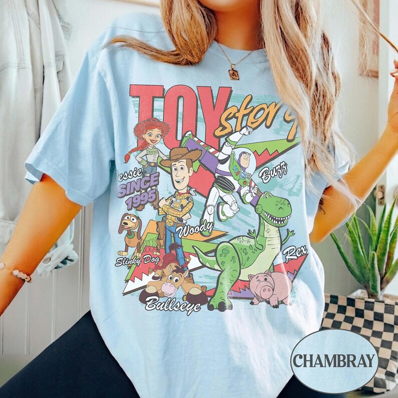 May include: A light blue t-shirt featuring a graphic print of Toy Story characters including Woody, Buzz Lightyear, Jessie, Rex, Slinky Dog, and Bullseye. The design includes the text Toy Story, Since 1995, and the names of the characters in various fonts.