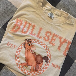 Pu&ograve; includere: T-shirt color crema con la scritta "BULLSEYE" in lettere maiuscole color corallo sopra un'illustrazione di un cavallo dei cartoni animati. Il cavallo &egrave; all'interno di un cerchio a scacchi color corallo e bianco, con "EST 1995" intorno. La maglietta &egrave; piegata su una superficie a scacchi.