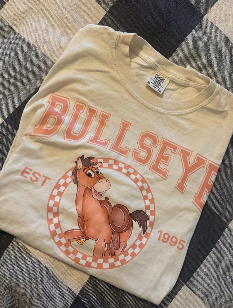Pu&ograve; includere: T-shirt color crema con la scritta "BULLSEYE" in lettere maiuscole color corallo sopra un'illustrazione di un cavallo dei cartoni animati. Il cavallo &egrave; all'interno di un cerchio a scacchi color corallo e bianco, con "EST 1995" intorno. La maglietta &egrave; piegata su una superficie a scacchi.