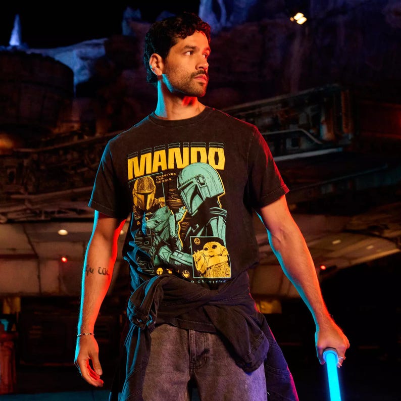 以下が含まれることがあります： 黄色いブロック体の「MANDO」の文字が入った黒いTシャツ。マンダロリアンとグローグを含む、スターウォーズシリーズのキャラクターのイラストが描かれています。画像内の人物は青いライトセーバーを持っています。