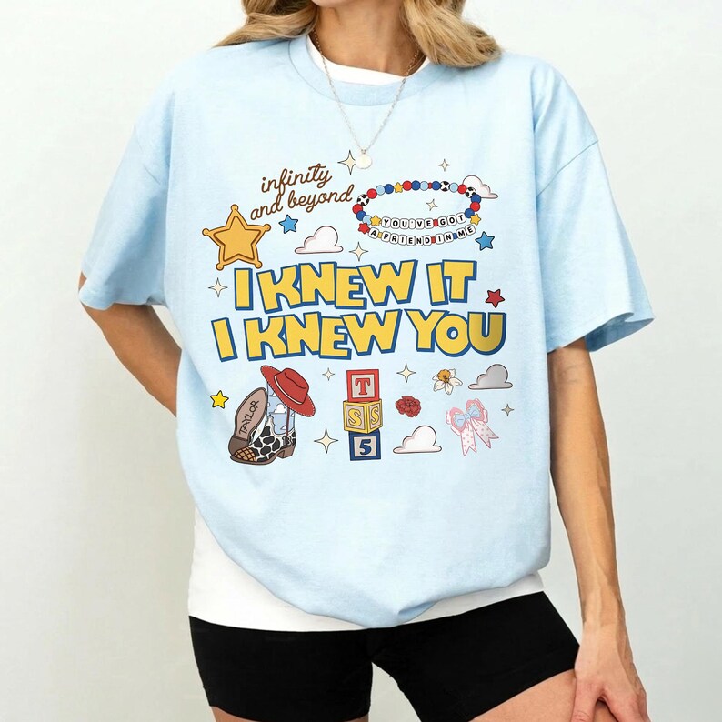 May include: A light blue t-shirt featuring a graphic print with the text "infinity and beyond", "YOU'VE GOT A FRIEND IN ME", and "I KNEW IT I KNEW YOU". The design includes a yellow star, a beaded bracelet, a cowboy boot, alphabet blocks, and a bow.