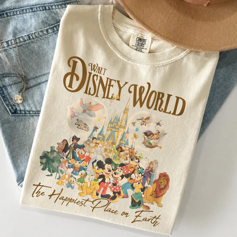 Op de afbeelding: Cr&egrave;mekleurig T-shirt met de tekst "Walt Disney World" in bruin script boven een kleurrijke afbeelding van Disney-personages en het kasteel van Assepoester. De zin "The Happiest Place on Earth" staat eronder.