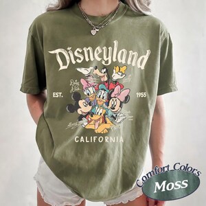 May include: Olive green t-shirt featuring the Disneyland logo and various Disney characters. The shirt includes the text "EST. 1955" and "CALIFORNIA". The shirt is a Comfort Colors Moss color.