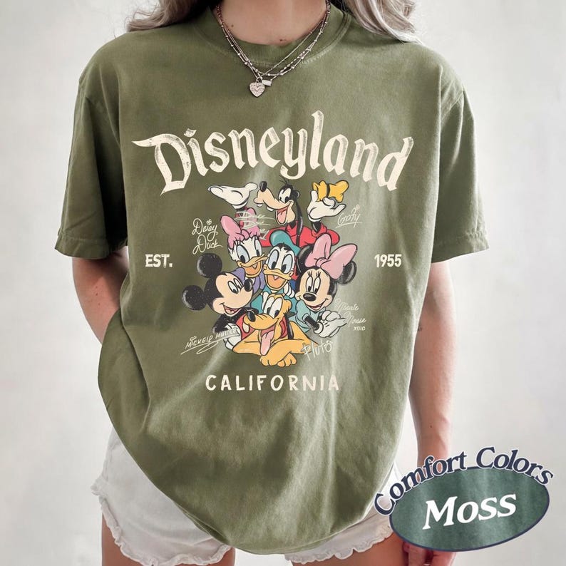May include: Olive green t-shirt featuring the Disneyland logo and various Disney characters. The shirt includes the text "EST. 1955" and "CALIFORNIA". The shirt is a Comfort Colors Moss color.