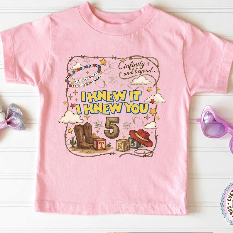 May include: A light pink short-sleeve t-shirt featuring a graphic print with the text "I KNEW IT I KNEW YOU", "Cinfinity and beyond", and "YOU'VE GOT A FRIEND IN ME". The design includes cowboy boots, a hat, blocks, and stars.