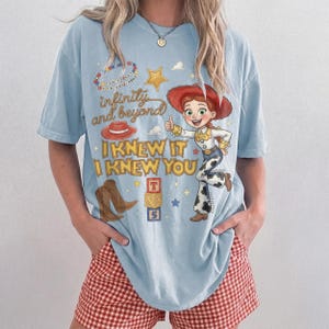 Może przedstawiać: Jasnoniebieski t-shirt z kr&oacute;tkim rękawem z nadrukiem graficznym przedstawiającym postać kowbojki, czerwony kapelusz, brązowe buty i klocki z literami. Tekst głosi "infinity and beyond", "I KNEW IT I KNEW YOU" oraz "YOU'VE GOT A FRIEND IN ME".
