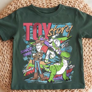 May include: Dark green t-shirt featuring the words "Toy Story" in a colorful, distressed font. The shirt displays Woody, Buzz Lightyear, and Rex the dinosaur. The text "Since 1995" is also included.