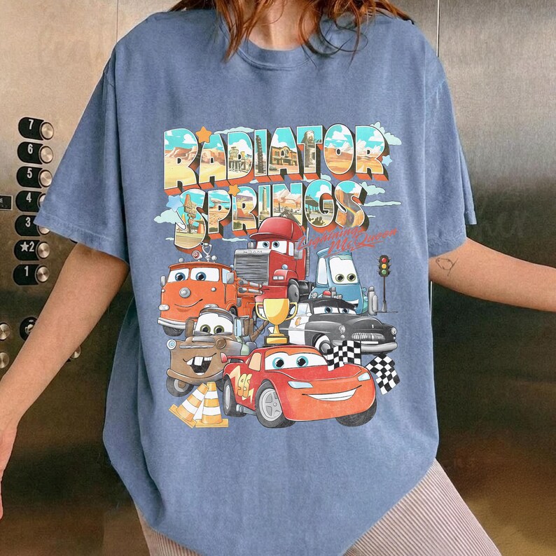 May include: Blue t-shirt featuring a colorful graphic of various cartoon cars with the text "RADIATOR SPRINGS" in a retro font. The design includes a desert landscape and a checkered flag.