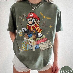 May include: Moss green t-shirt featuring a cartoon Mario character dressed as a wizard, sitting on books. The design includes a wand, glasses, a scarf, and a wizard hat. The shirt has the word "MOSS" in a circle on the bottom right.