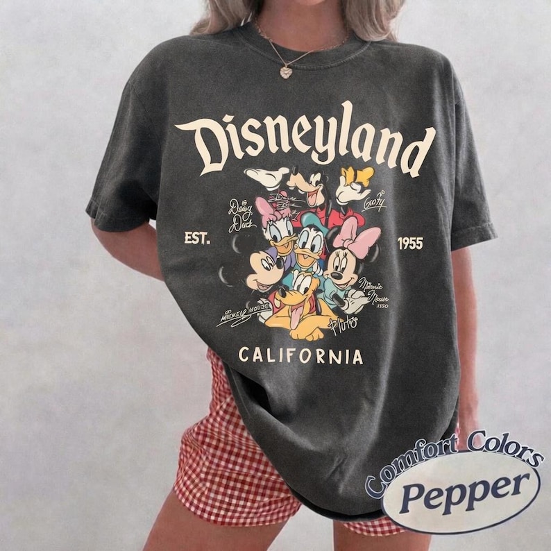 May include: Dark gray t-shirt featuring the Disneyland logo and various Disney characters. The shirt includes the text "EST. 1955" and "CALIFORNIA". The shirt is a Comfort Colors Pepper brand.
