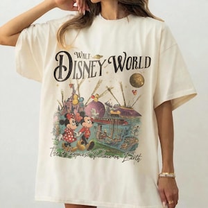 May include: Cream-colored t-shirt featuring a vintage Walt Disney World graphic. The design includes Mickey and Minnie Mouse, a castle, and the text "The Happiest Place on Earth". The shirt has short sleeves and a relaxed fit.