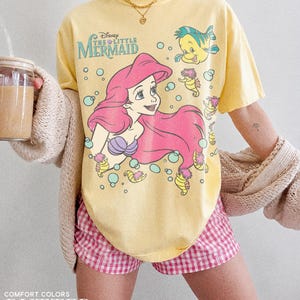 May include: A butter yellow short-sleeve t-shirt featuring a graphic print of Ariel from The Little Mermaid with pink hair, a purple seashell top, Flounder, and seahorses surrounded by teal bubbles. The text Disney The Little Mermaid is printed.