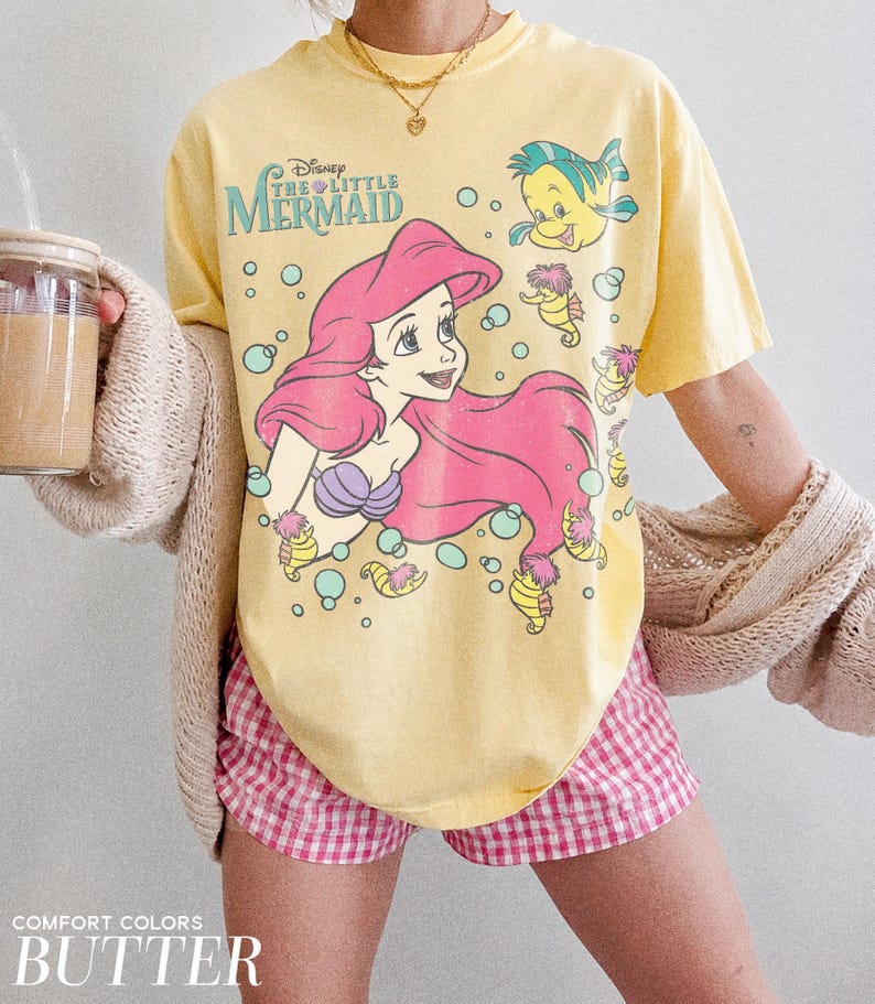 May include: A butter yellow short-sleeve t-shirt featuring a graphic print of Ariel from The Little Mermaid with pink hair, a purple seashell top, Flounder, and seahorses surrounded by teal bubbles. The text Disney The Little Mermaid is printed.