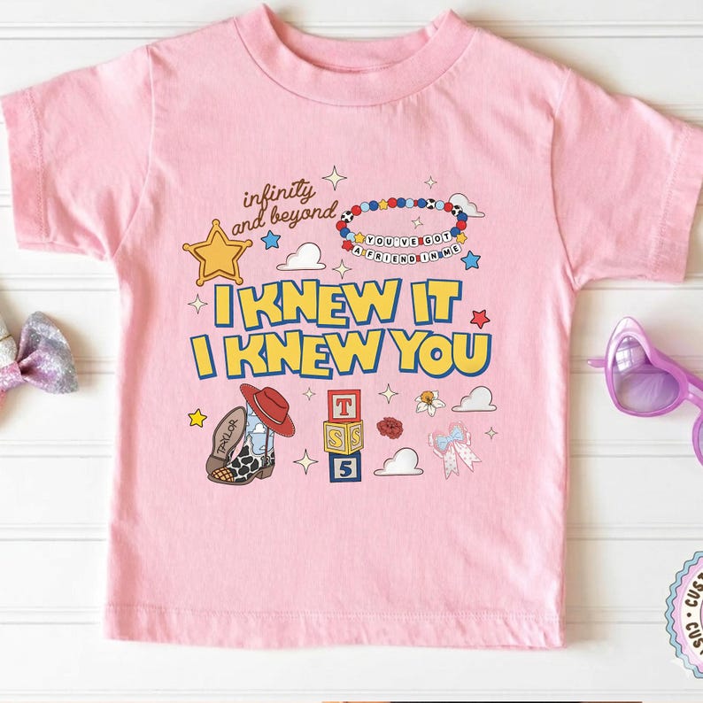 May include: A light pink short-sleeve t-shirt featuring a graphic print with the text "infinity and beyond", "I KNEW IT I KNEW YOU", and "YOU'VE GOT A FRIEND IN ME". The design includes a yellow star, a beaded bracelet, a cowboy boot, and alphabet blocks.