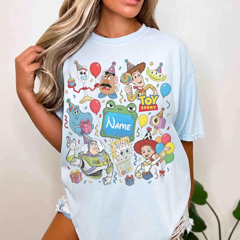 May include: A light blue t-shirt featuring a colorful graphic print of various Toy Story characters wearing party hats, including Woody, Buzz Lightyear, Jessie, and Forky, with balloons, gifts, and a central green frame with the text Name.