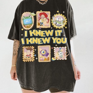 May include: A black oversized t-shirt featuring a graphic print of Toy Story characters in postage stamp frames. The text reads I KNEW IT I KNEW YOU. The design includes Buzz Lightyear, Woody, Jessie, and other characters, along with small stars and a ball.