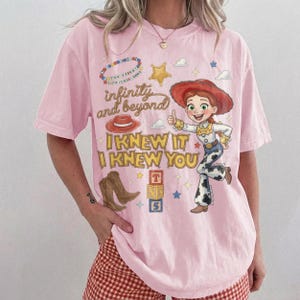 Może przedstawiać: Jasnor&oacute;żowy t-shirt z kr&oacute;tkim rękawem z nadrukiem graficznym przedstawiającym kowbojkę, czerwony kapelusz, brązowe buty i klocki z literami. Tekst głosi infinity and beyond, I KNEW IT oraz I KNEW YOU, w otoczeniu gwiazd i chmur.
