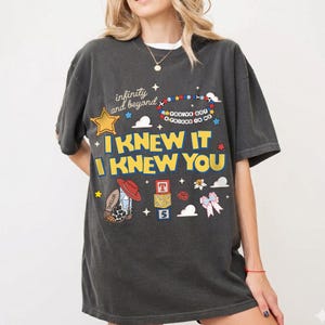 May include: A charcoal gray short-sleeve t-shirt featuring a graphic print with the text "infinity and beyond", "YOU'VE GOT A FRIEND IN ME", and "I KNEW IT I KNEW YOU". The design includes a yellow star, a beaded bracelet, a cowboy boot, blocks, and a bow.