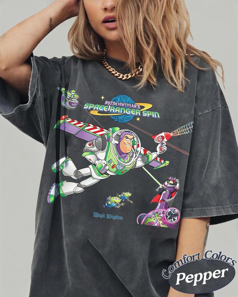 May include: A charcoal gray short-sleeve t-shirt featuring a graphic of Buzz Lightyear from Space Ranger Spin. The design includes Buzz Lightyear flying, green aliens, Zurg, and the text Buzz Lightyear's Space Ranger Spin and Magic Kingdom.