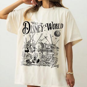 May include: Cream-colored t-shirt featuring a black and white graphic of Mickey and Minnie Mouse in front of a Walt Disney World scene. The text "Walt Disney World" and "The Happiest Place on Earth" are also printed on the shirt.